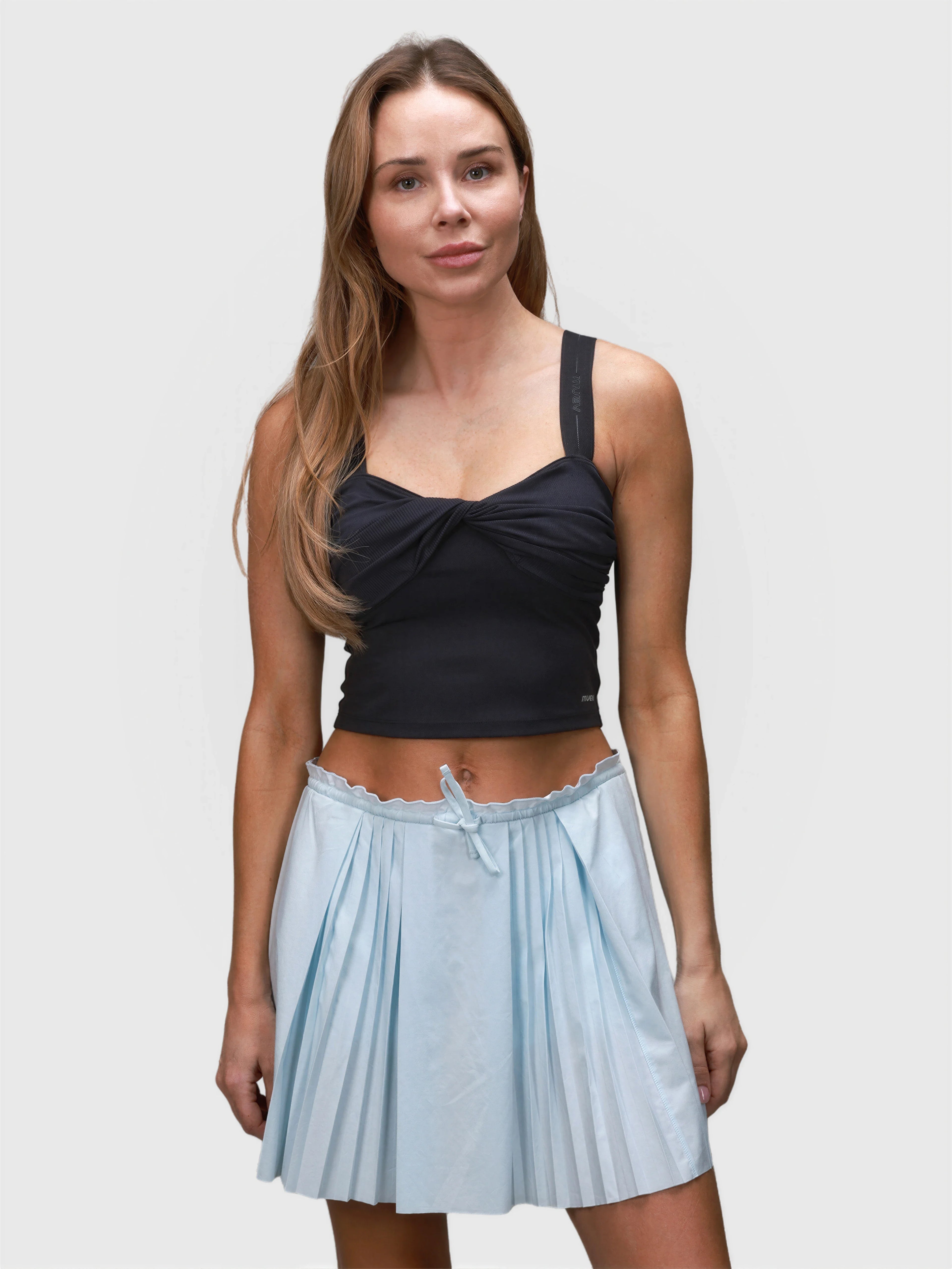 Front view of woman wearing black Airy Twist Tank by MUEV paired with light blue pleated skirt for padel apparel